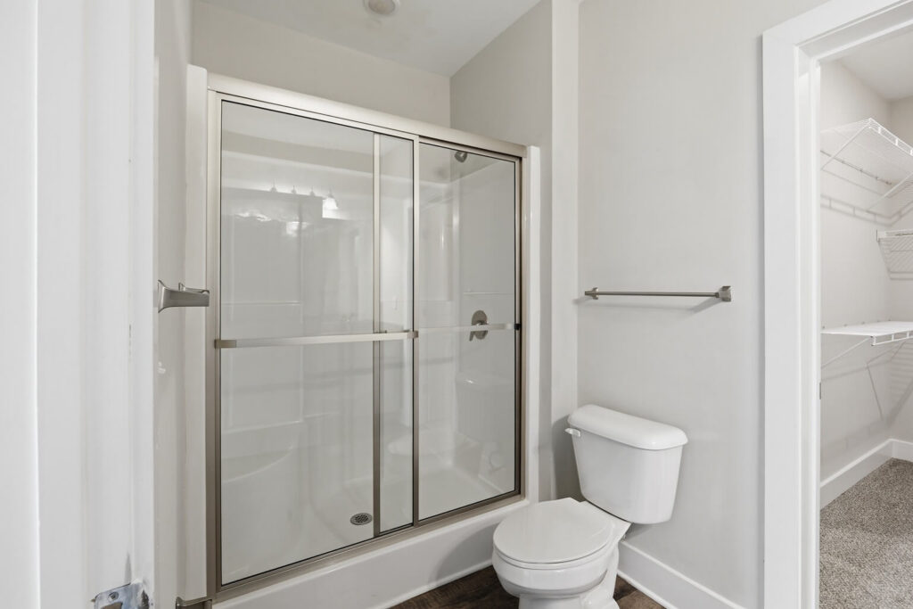 A modern bathroom with a glass-enclosed shower, white toilet, towel bar, and an adjacent walk-in closet with wire shelving. The walls are light-colored and the flooring is dark wood.