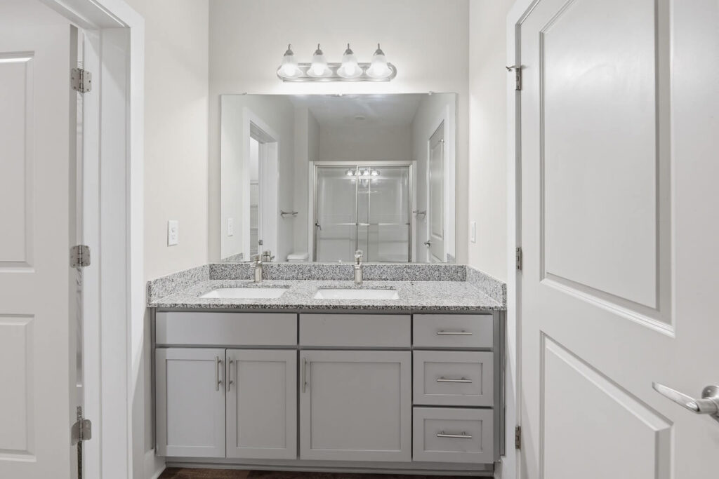 Modern bathroom with a double sink vanity, granite countertop, large mirror, silver fixtures, and white cabinets. Four lights are above the mirror, and doors are visible on both sides. The overall color scheme is light and neutral.