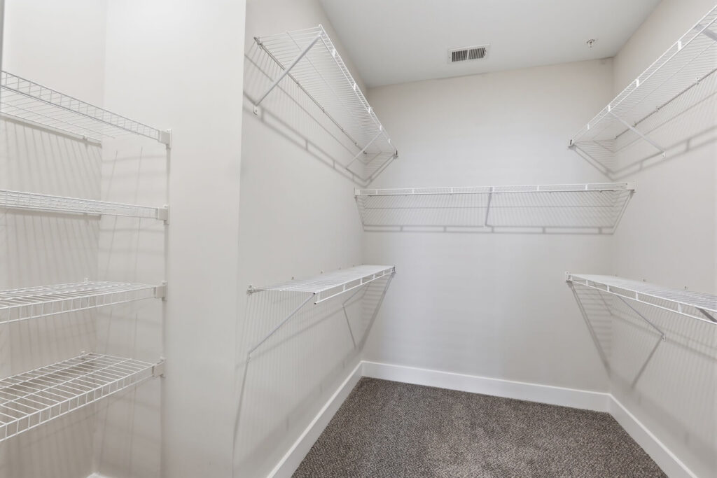 Empty walk-in closet with white wire shelving on both sides and back wall, beige carpeted floor, and light-colored walls. Ceiling vent is visible near the back wall.