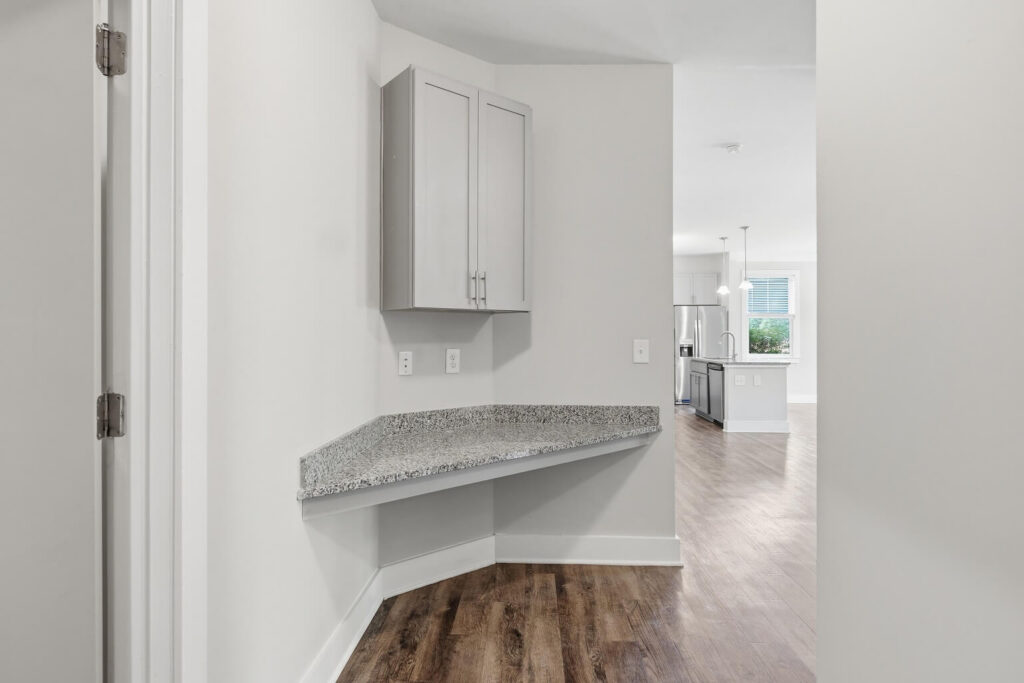 A small nook with a granite corner desk, upper cabinets, and outlets; it opens to a bright kitchen with wood floors, white cabinets, and stainless steel appliances.