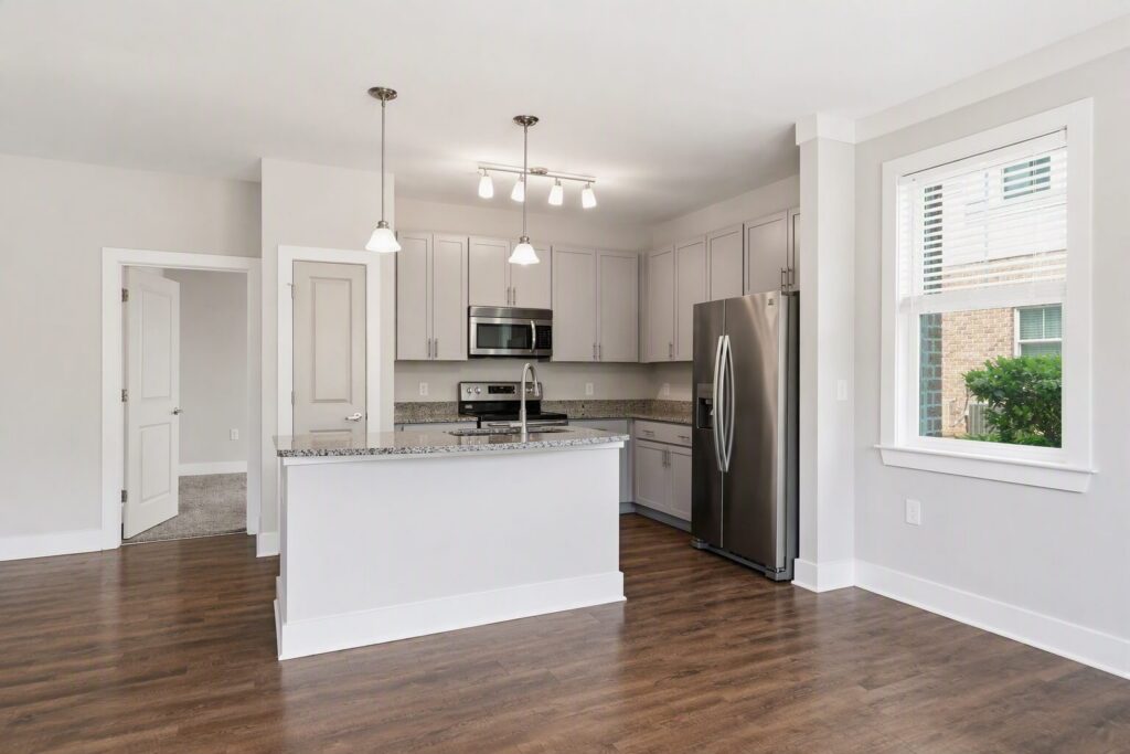 Modern kitchen with white cabinets, stainless steel appliances, granite countertops, pendant and ceiling lights, hardwood floor, and a window letting in natural light. A door leads to an adjacent room.